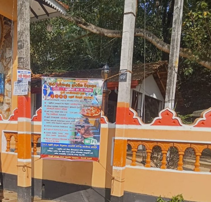 Enrollment banner tied between poles beside an orange temple wall on a road in Dampella